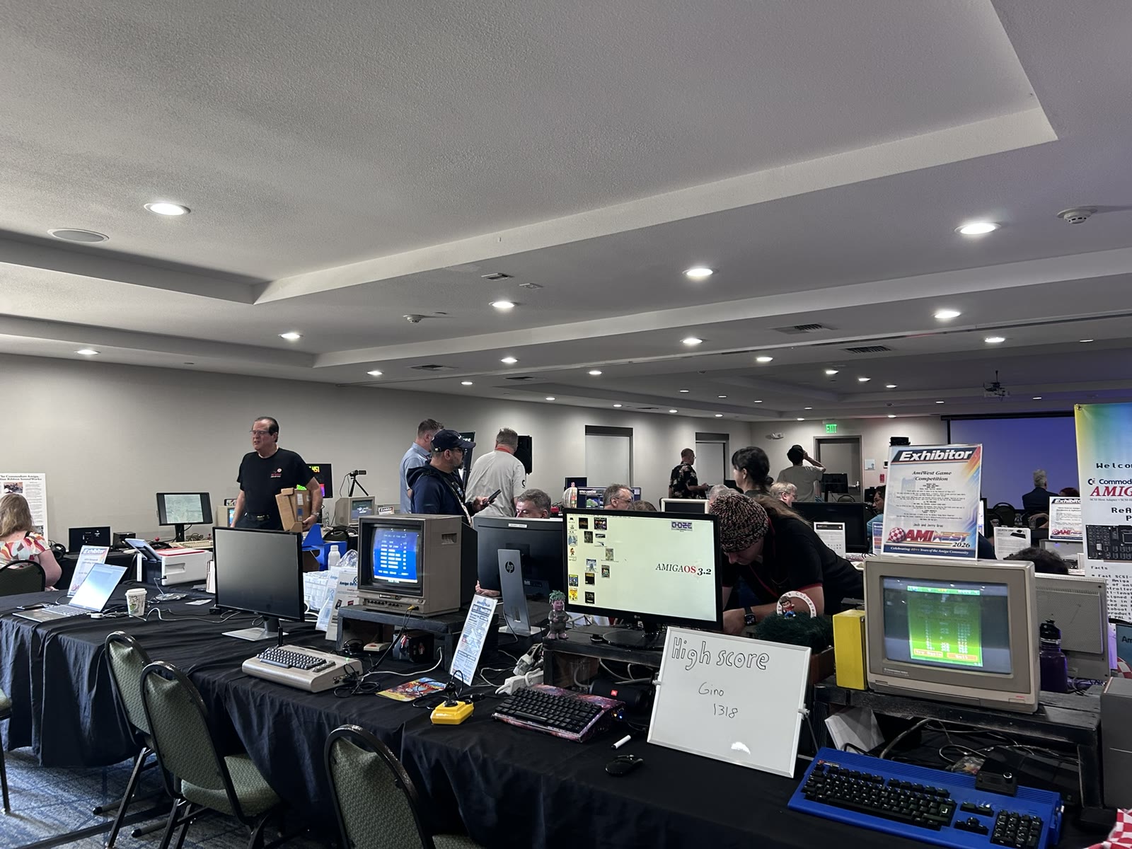 AmiWest exhibition tables with CRT displays, an AmigaOS 3.2 screen, keyboards and a game competition sign